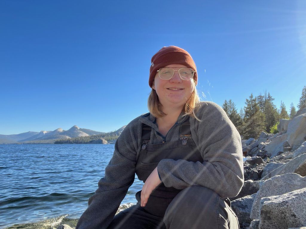 The image shows a person wearing glasses and a beanie, sitting on rocks near a lake. The person is smiling and appears to be enjoying the sunny day. In the background, there are mountains and trees, with a clear blue sky overhead.